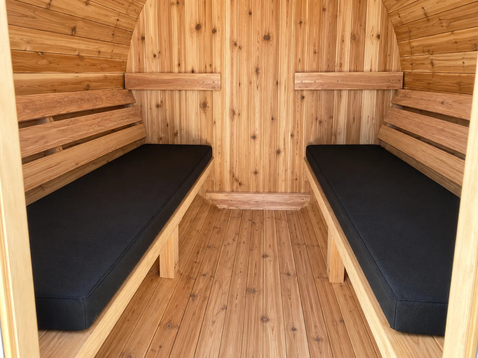 Two fitted black sauna cushions on facing benches inside a cedar barrel sauna